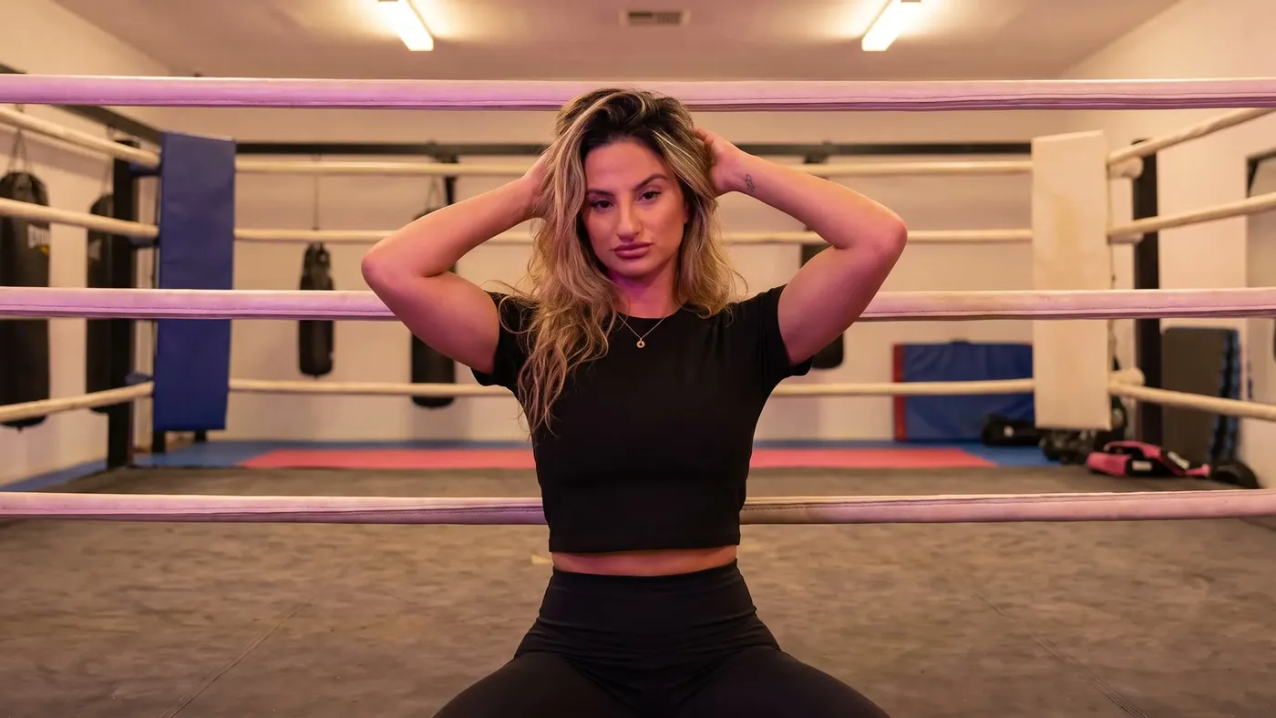 Jana sitting on the gym floor against a boxing ring with her hands in her hair, relaxed after training
