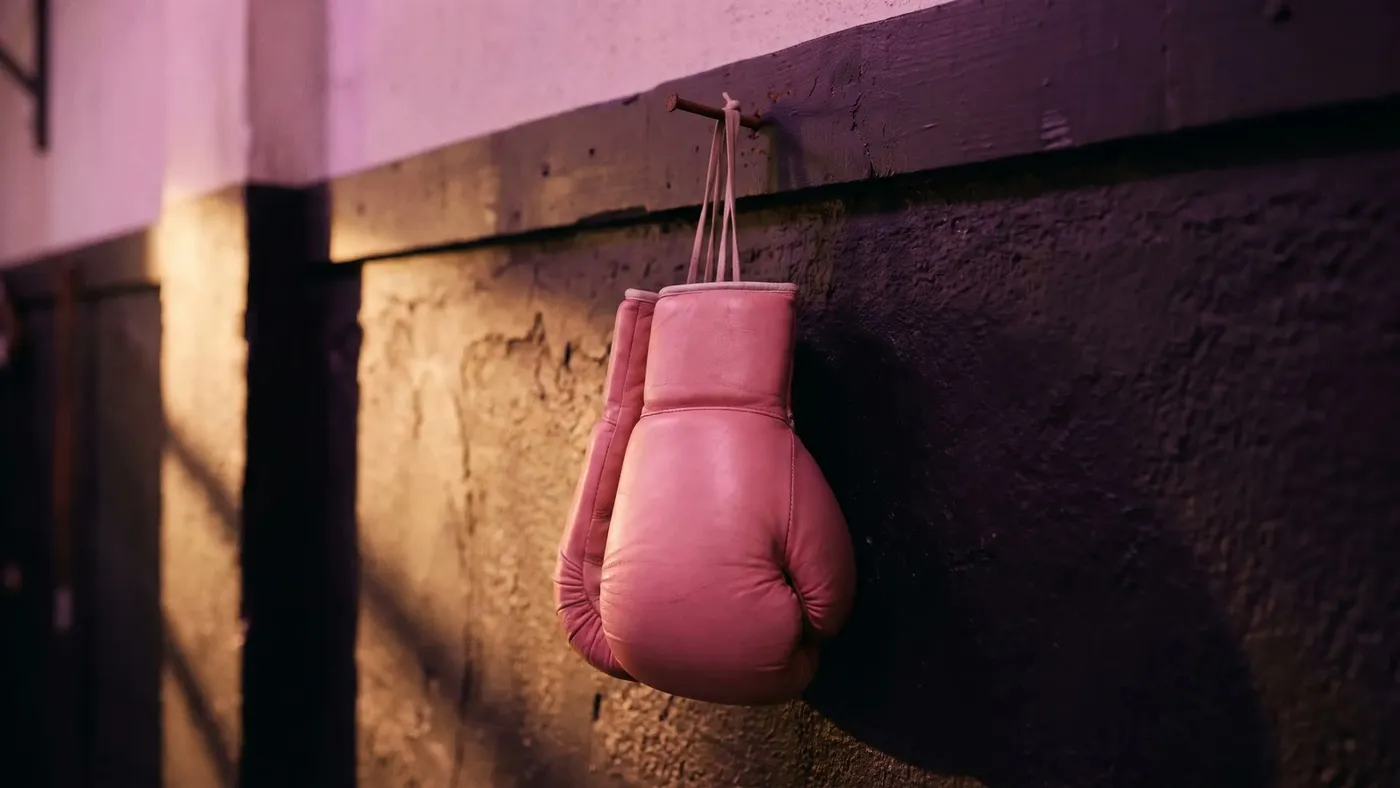 A pair of pink boxing gloves hanging from a nail on a dark gym wall in warm light