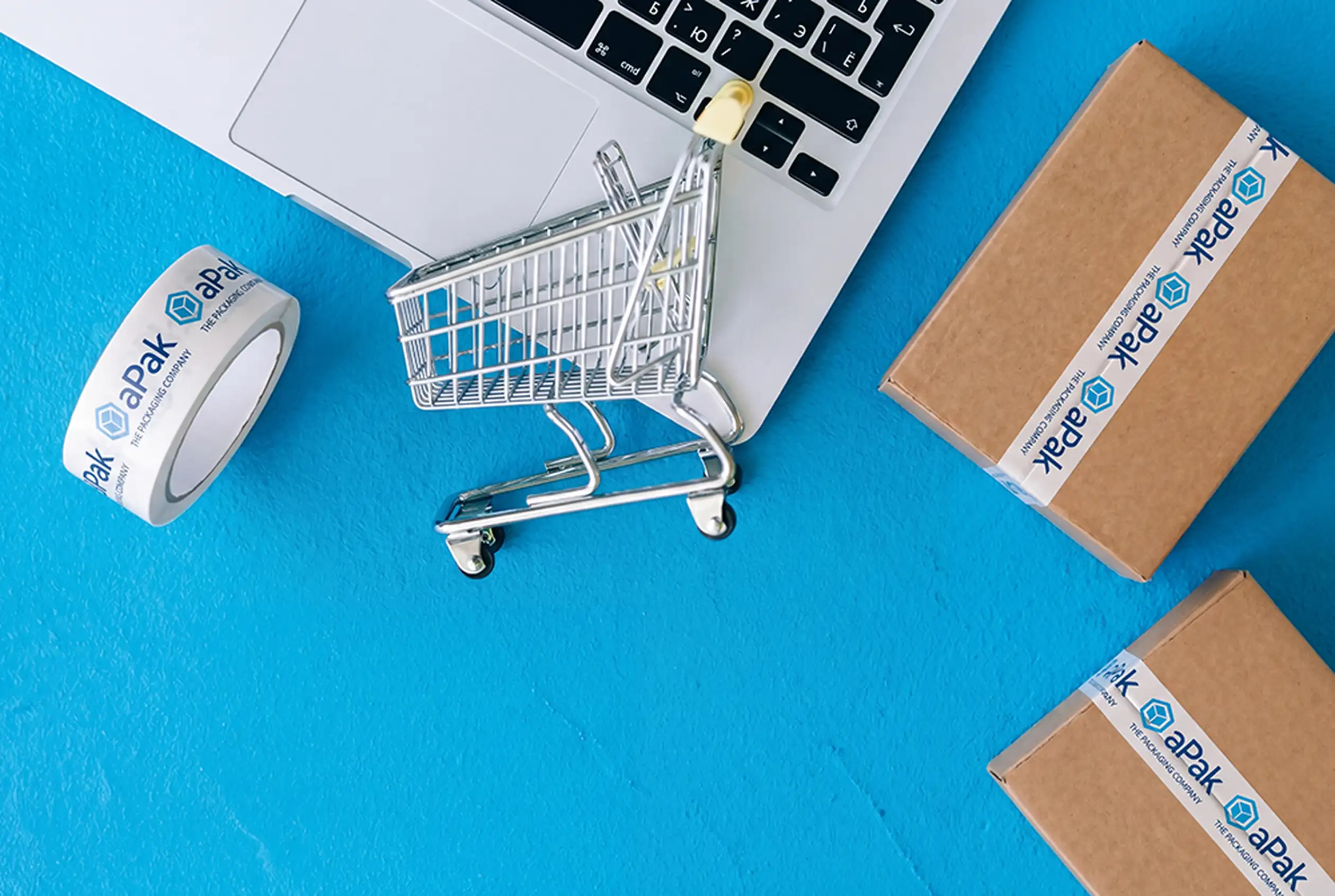 A laptop, mini shopping cart, roll of branded tape, and cardboard boxes on a blue surface, representing online shopping and packaging.