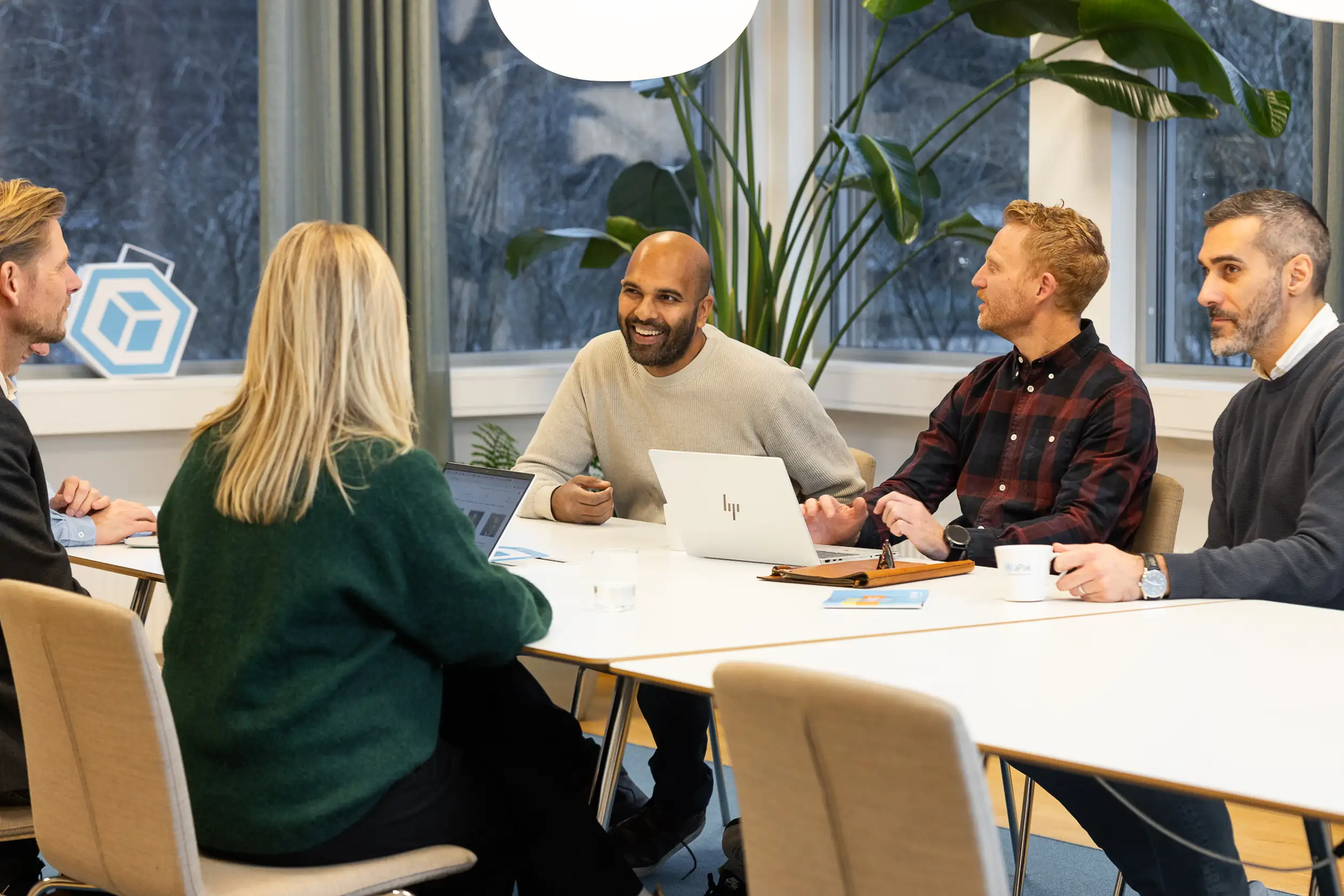 Five people sitting around a conference table, engaged in a meeting, with laptops and coffee cups in a modern office setting.