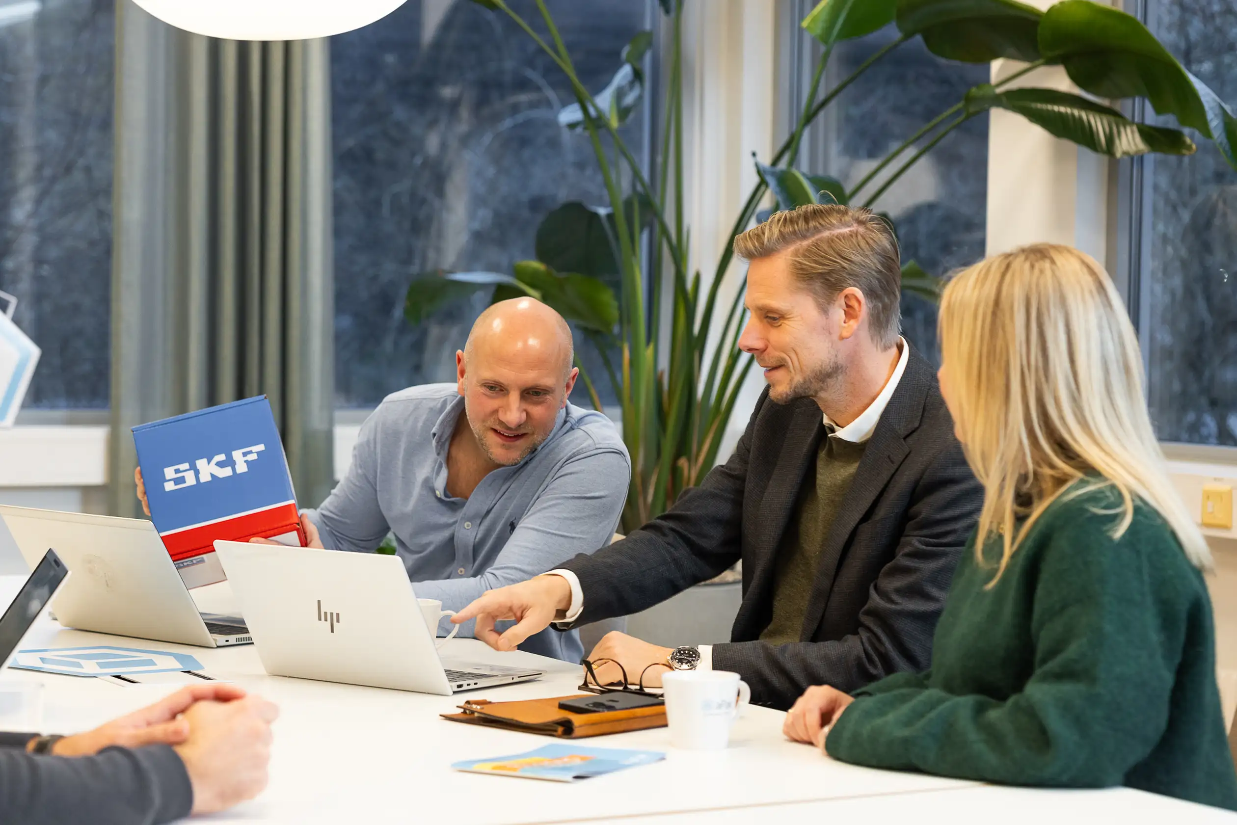 Three people in an office setting engaged in discussion, with laptops open and a visible SKF logo on a box.