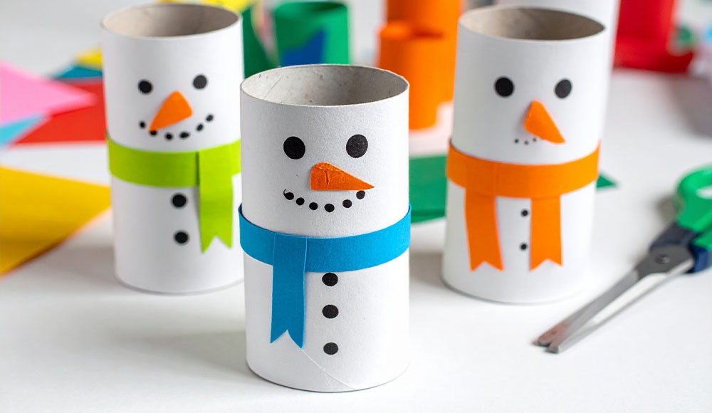 Klopapier-Schneemänner Gebastelte Klopapier-Schneemänner mit bunten Schals aus Papier stehen auf einem hellen Tisch mit Bastelutensilien