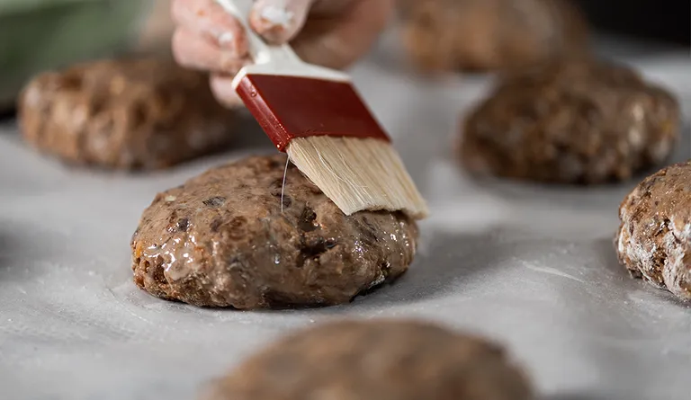 Mehrere frisch geformte Früchtebrote liegen auf einem großen Backblech und werden mit einem Pinsel bestrichen
