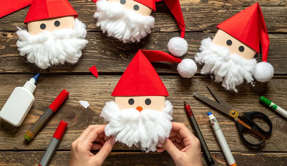 Weihnachtsmänner mit Filzbärtcen und Papiermützen auf Holztisch mit Bastelutensilien