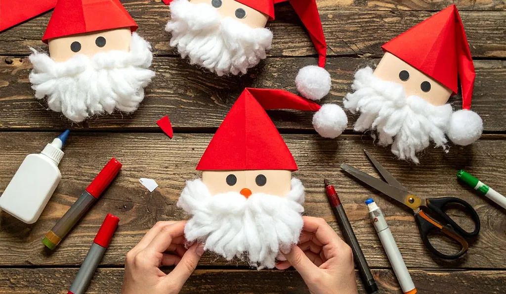 Weihnachtsmänner mit Filzbärtcen und Papiermützen auf Holztisch mit Bastelutensilien