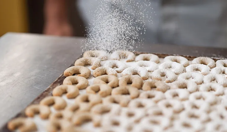 Frisch gebackene Vanillekipferl liegen auf einem Blech und werden mit Staubzucker bestreut