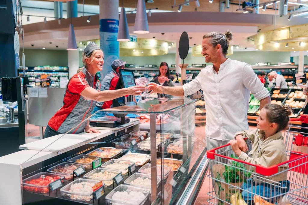 Ein Mann mit einem Kind im Einkaufswagen unterhält sich in einem Supermarkt mit einem lächelnden Mitarbeiter der Feinkostabteilung, umgeben von verschiedenen Lebensmitteln in den Auslagen.
