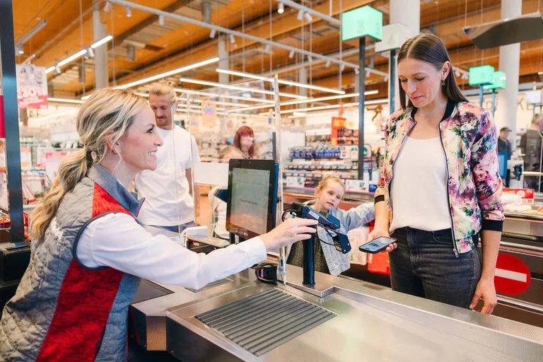 Eine Frau, die an der Kasse eines Supermarkts mit ihrem Smartphone bezahlt, während im Hintergrund eine lächelnde Kassiererin und ein neugierig zuschauendes Kind zu sehen sind.