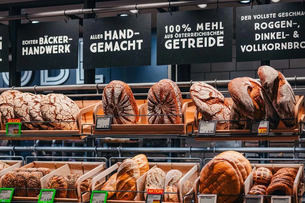 Verschiedene handgemachte Brotsorten, ausgestellt in den Regalen einer Bäckerei, mit Schildern, die auf österreichisches Getreide und handwerkliche Herstellung hinweisen.
