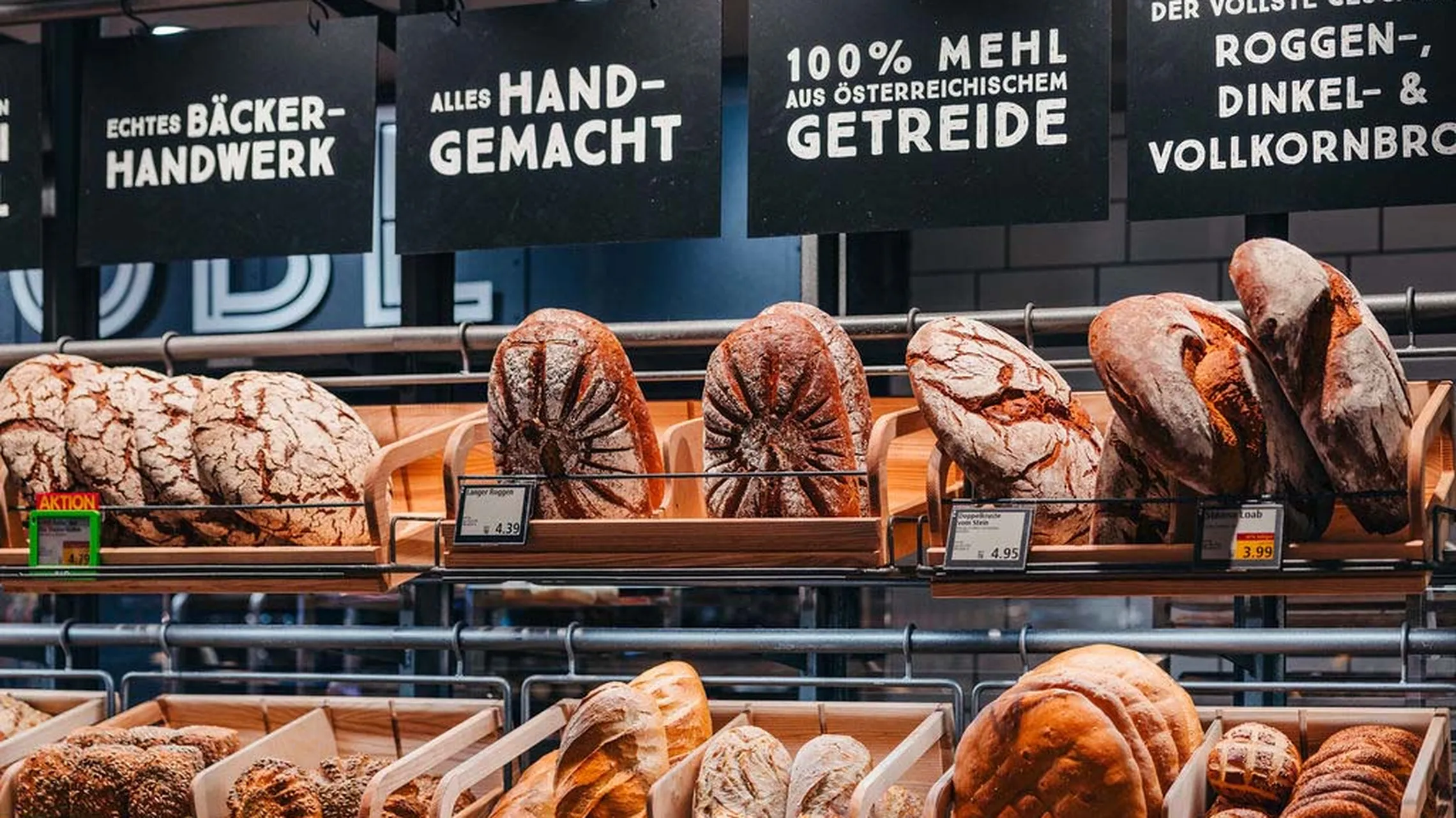 Verschiedene handgemachte Brotsorten, ausgestellt in den Regalen einer Bäckerei, mit Schildern, die auf österreichisches Getreide und handwerkliche Herstellung hinweisen.