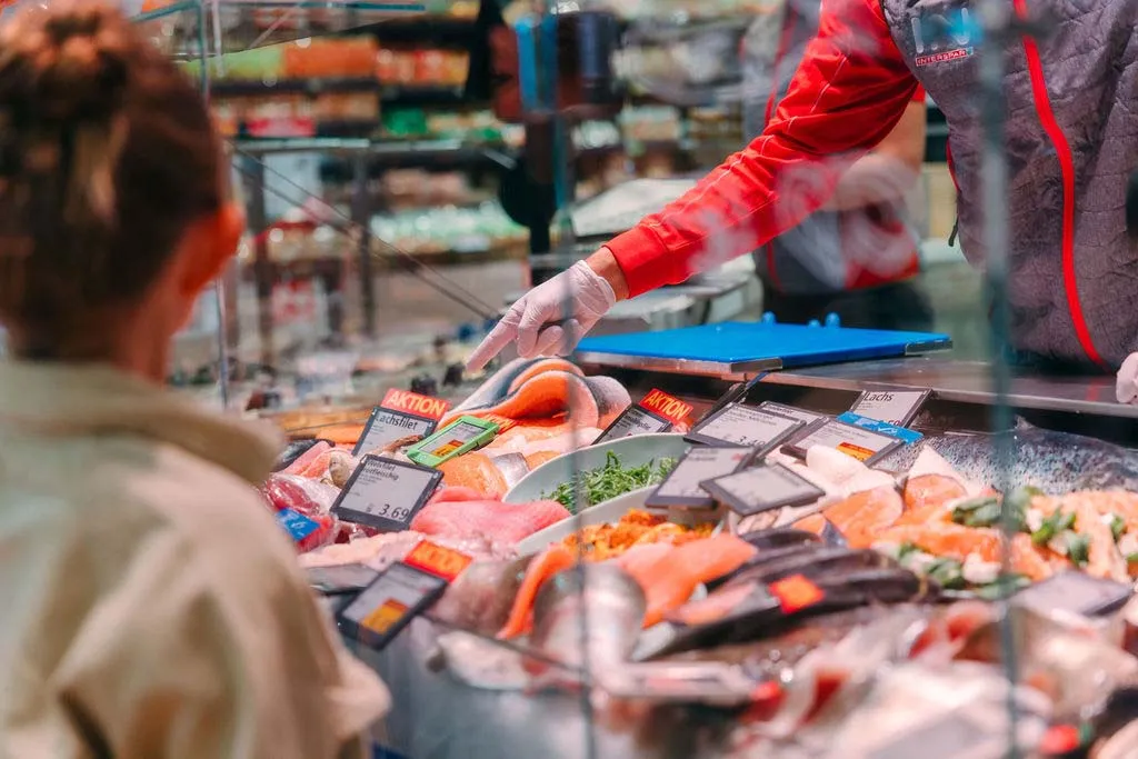 Eine Person sucht sich an der Fischtheke eines Supermarkts Fisch aus, wo verschiedene Stücke mit Preisschildern ausgestellt sind.