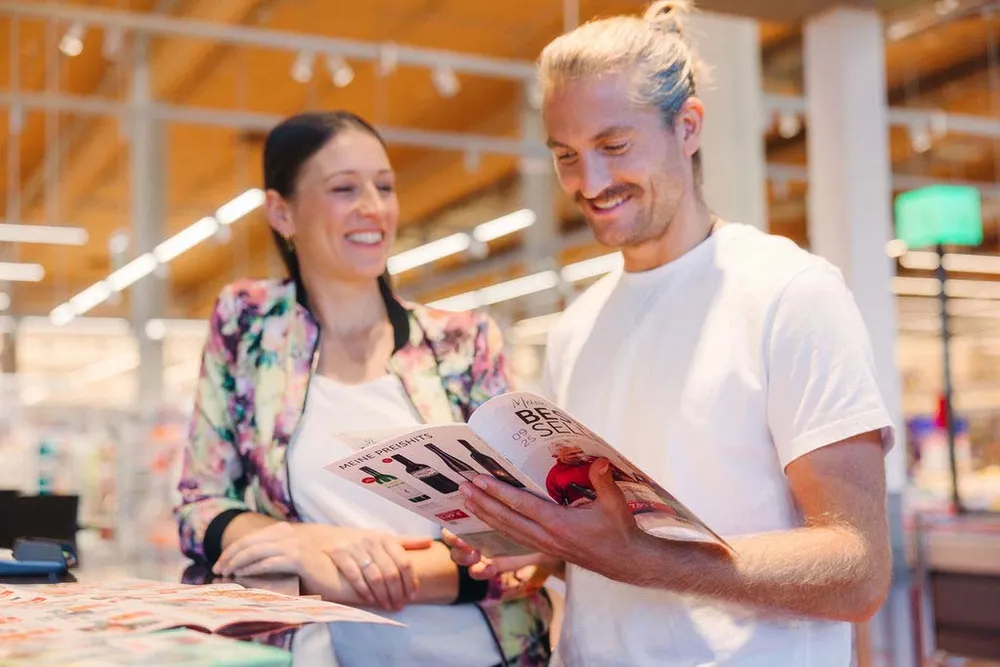 Ein lächelnder Mann und eine lächelnde Frau in einem Geschäft schauen sich gemeinsam einen Produktkatalog an, im Hintergrund sind helle Lichter und Regale zu sehen.
