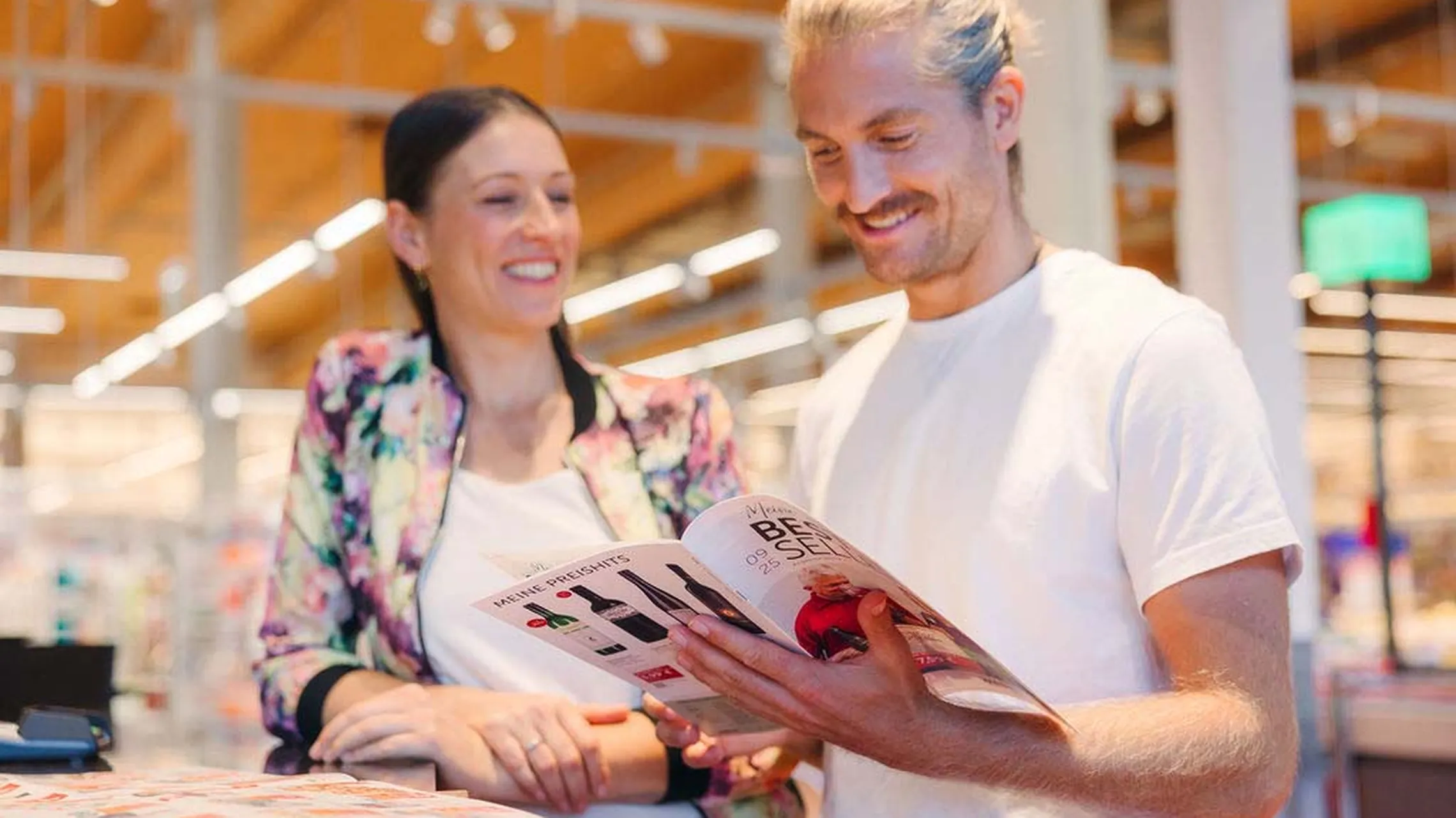 Ein lächelnder Mann und eine lächelnde Frau in einem Geschäft schauen sich gemeinsam einen Produktkatalog an, im Hintergrund sind helle Lichter und Regale zu sehen.