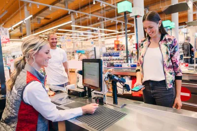 Eine Frau in einer geblümten Jacke bezahlt an der Kasse eines Supermarkts mit ihrem Handy und lächelt, während die Kassiererin ihr hilft. Hinter ihr steht ein Mann in der Schlange.