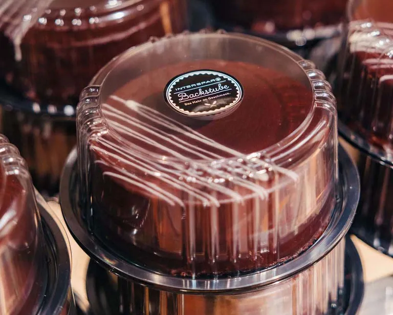 Ein Schokoladenkuchen in einem durchsichtigen, runden Plastikbehälter mit einem Etikett auf der Oberseite, der zwischen anderen ähnlich verpackten Kuchen steht.