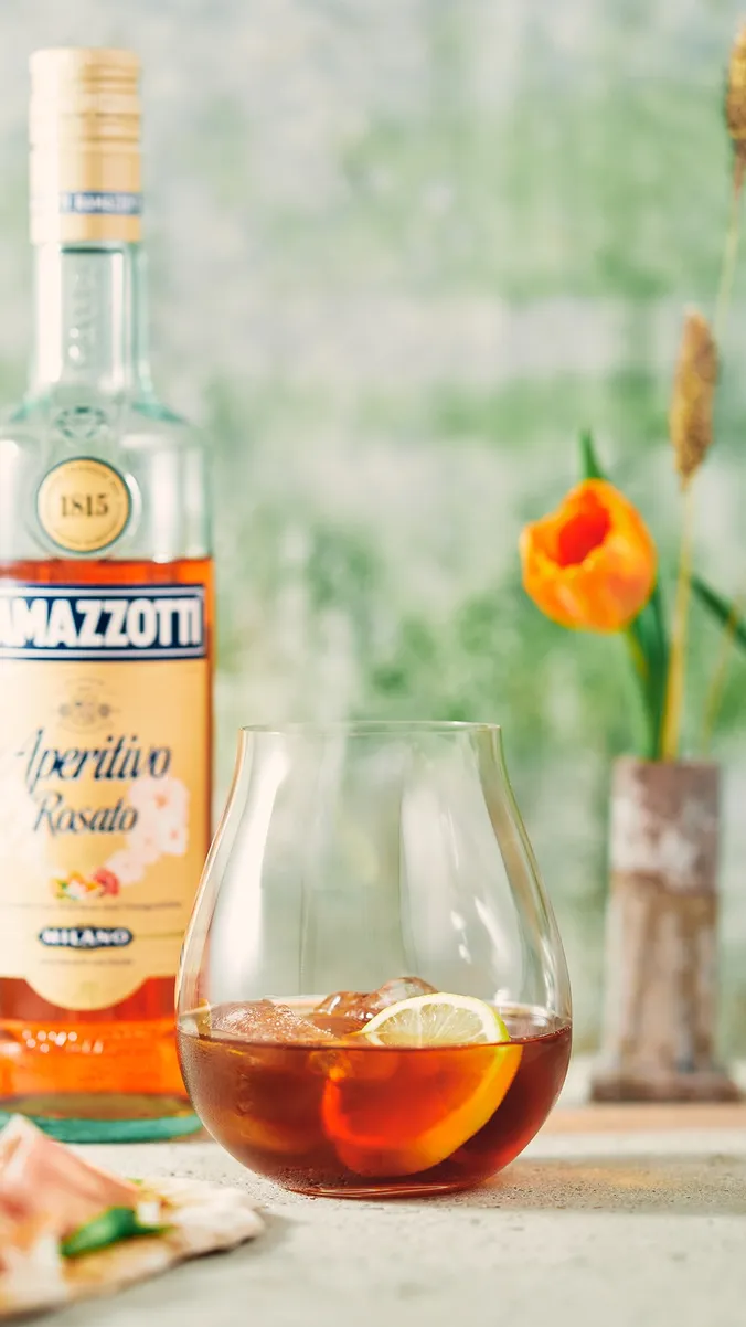 Ein Glas mit einem eisgekühlten, bernsteinfarbenen Getränk und einer Zitronenscheibe neben einer Flasche Aperitif; im Hintergrund eine orangefarbene Tulpe und Aufschnitt auf Brot.