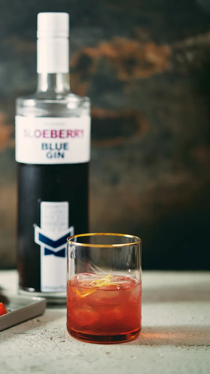 Ein Glas mit einem rötlichen Cocktail, garniert mit einer Zitronenschale, vor einer Flasche mit der Aufschrift „Sloeberry Blue Gin“ auf einer strukturierten Oberfläche.