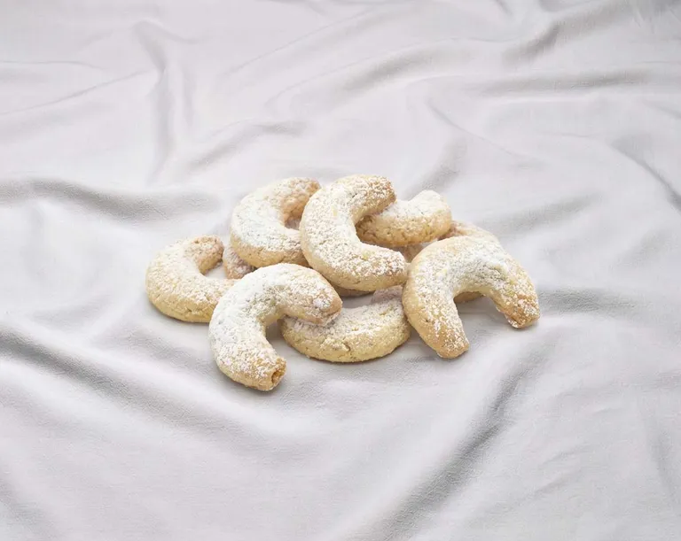 Ein Stapel halbmondförmiger Kekse, bestäubt mit Puderzucker, vor einem weichen, weißen Stoffhintergrund.