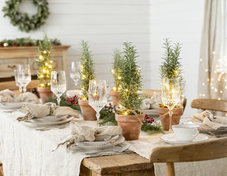 Ein Weihnachtstisch mit Leinentischdecke, edlem Porzellan, Weingläsern und Servietten, geschmückt mit Rosmarin-Topfpflanzen, die wie kleine Weihnachtsbäumchen mit Lichterketten verziert sind. Eine fröhliche, festliche Atmosphäre im Landhausstil.