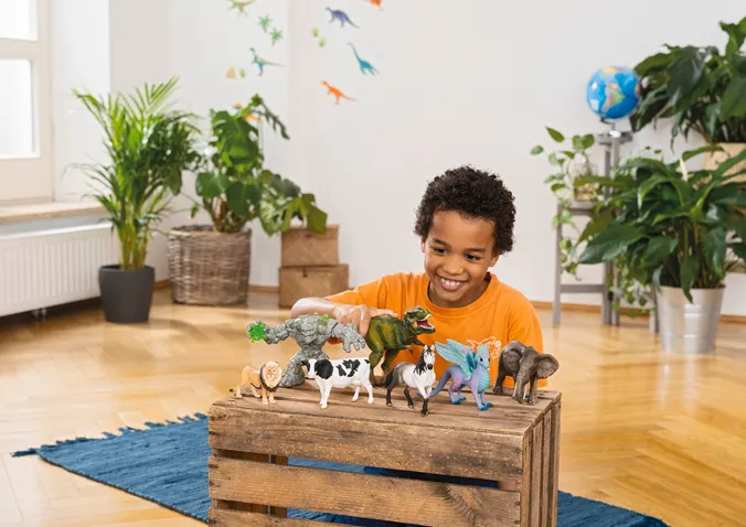 Junge in orangem T-Shirt spielt mit verschiedenen Spielfiguren am Boden, im Hintergrund Zimmerpflanzen