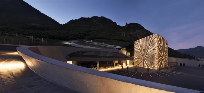 Ein modernes Gebäude mit einer geometrischen Fassade, die in der Abenddämmerung leuchtet, vor einer Bergkulisse und mit einer kurvenreichen Straße im Vordergrund.