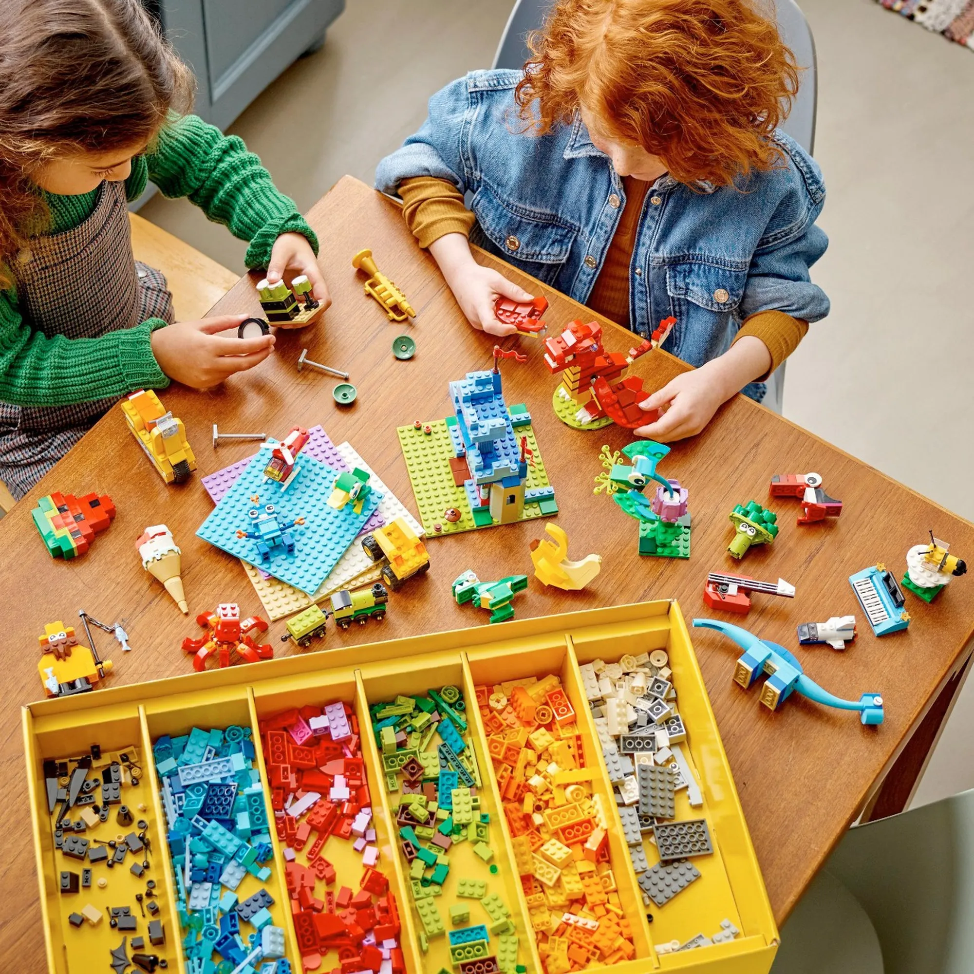 2 Mädchen spielen auf einem Holztisch mit Legos, Bauwerke sowie Steine sind am Tisch verteilt