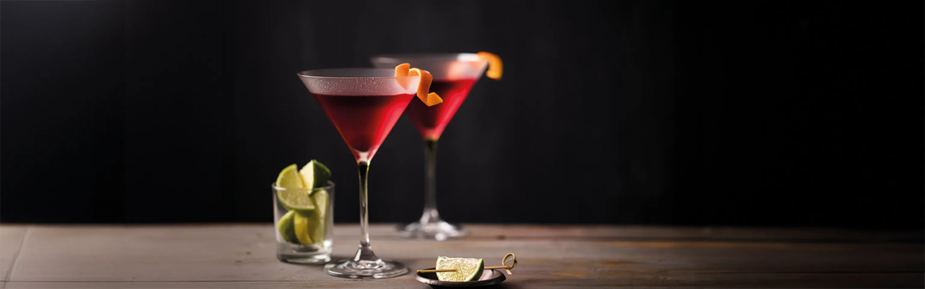 Zwei Cosmopolitan-Cocktails im Martini-Glas mit Orangenzeste und Limettenspalten