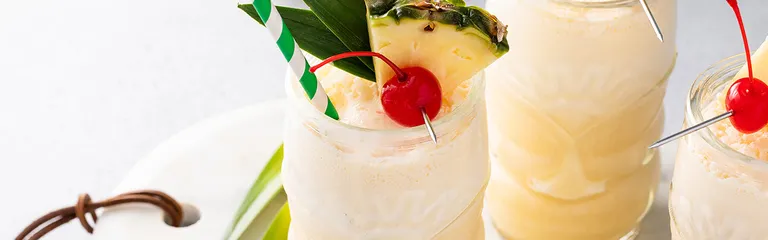 Drei Gläser mit Pina Colada in Tiki-Gläsern. Darin ein Stück Ananas, eine Amarenakirsche und ein Strohhalm, sowie Palmenblätter.