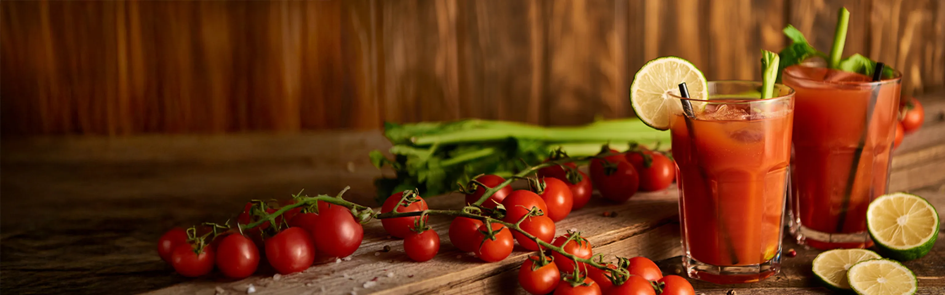 Zwei Bloody-Mary-Cocktails mit Limette und Sellerie neben frischen Tomaten auf einem Holztisch
