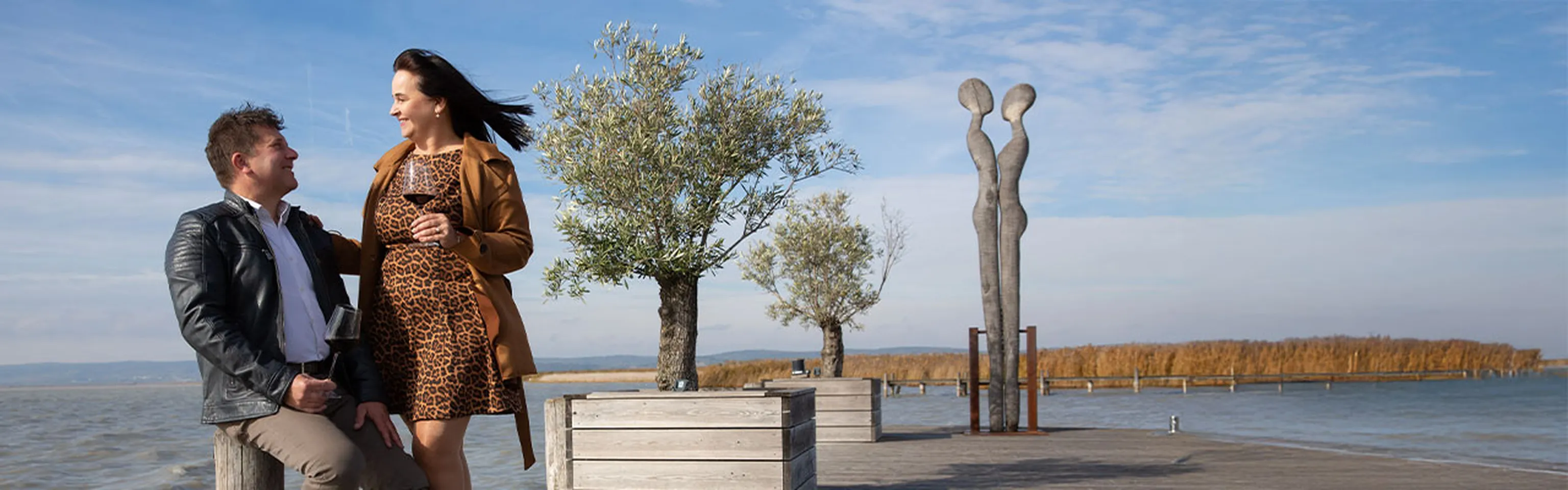 Robert und Marietta Keringer am Neusiedlersee, im Hintergrund zwei Figuren aus Holz, sowie zwei kleine Bäume.
