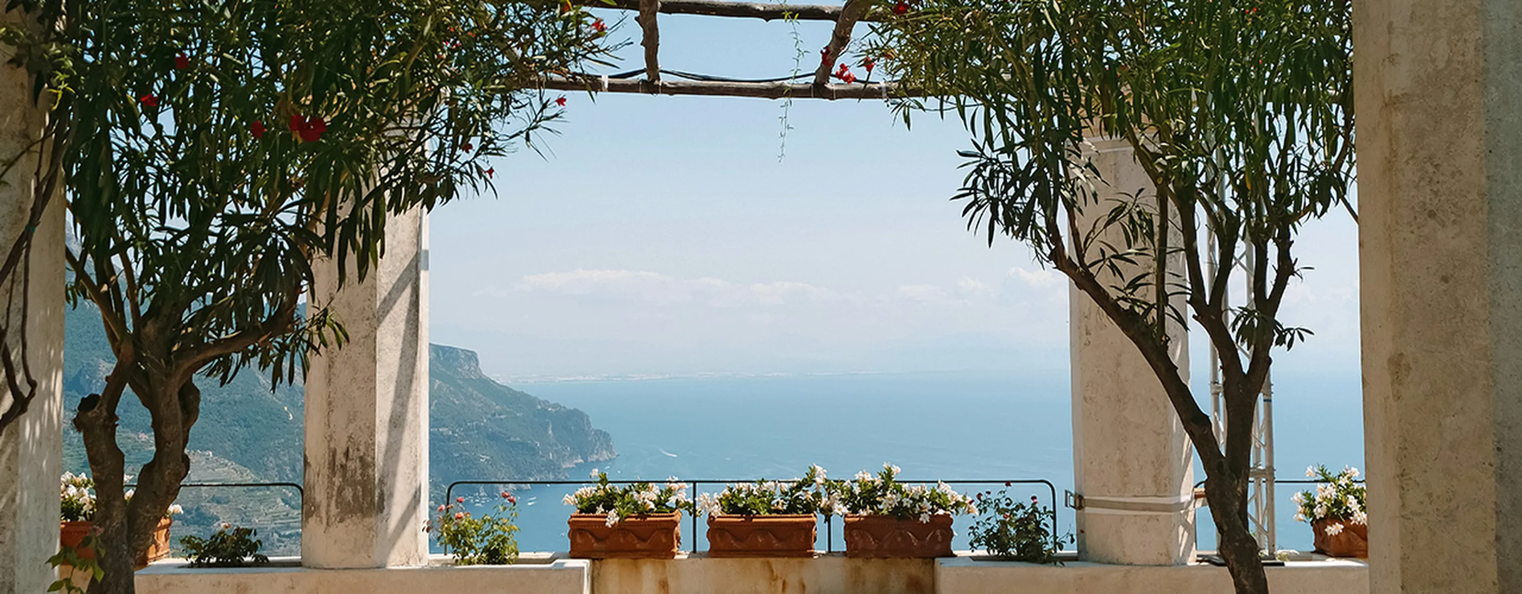 A scenic view through an archway with trees and flowers, overlooking a coastline and ocean under a clear blue sky.
