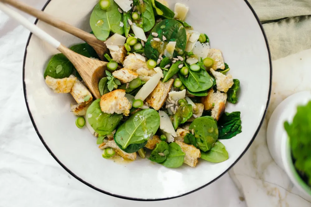 Frischer Spinatsalat mit Croutons, Spargel, Käsespänen und Samen in einer Schüssel, mit Holzbesteck auf einer weißen Tischdecke angerichtet..