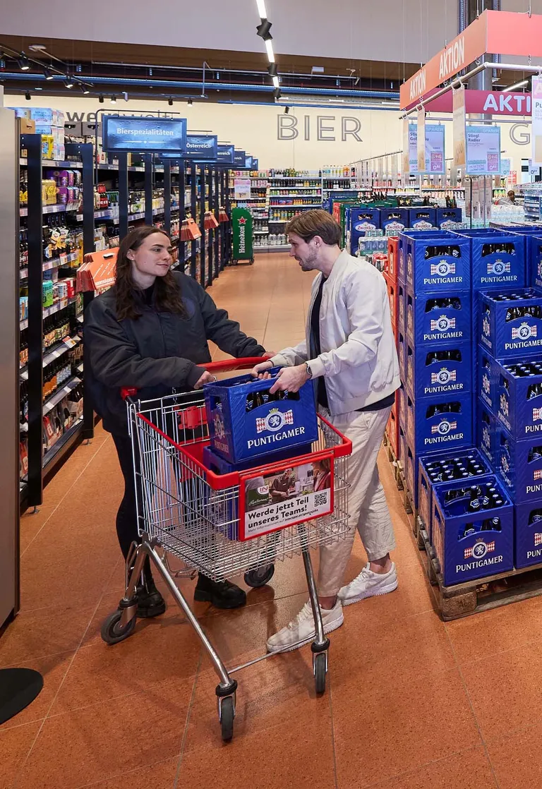 Zwei Personen in einem Supermarkt, die eine Kiste Bier in einen Einkaufswagen legen. Um sie herum stehen Regale mit Getränken und Verkaufsschilder.