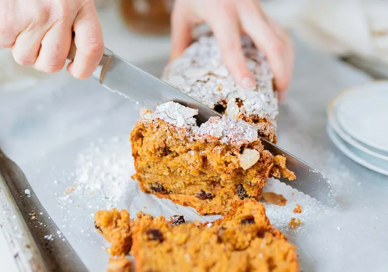 Auf einem mit Backpapier ausgelegten Blech schneiden Hände ein mit Puderzucker bestreutes Rührkuchenstück mit Rosinen und Nüssen in Scheiben.