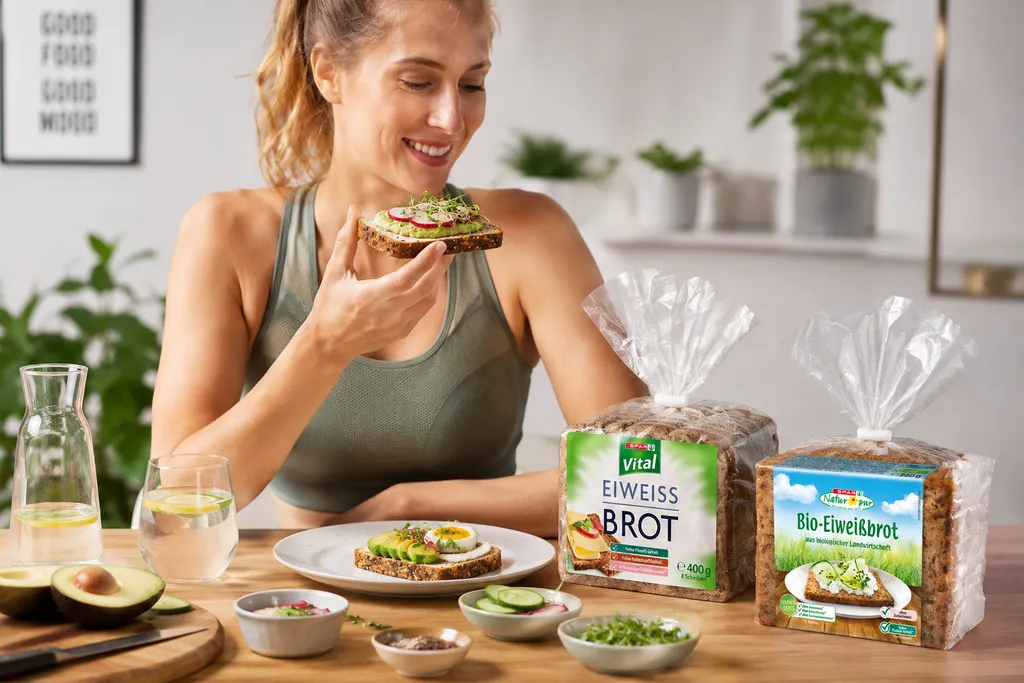 Frau genießt ein belegtes Eiweißbrot mit Avocado und frischem Gemüse. Daneben stehen SPAR Vital Eiweißbrot und SPAR Natur*pur Bio-Eiweißbrot auf einem Frühstückstisch.