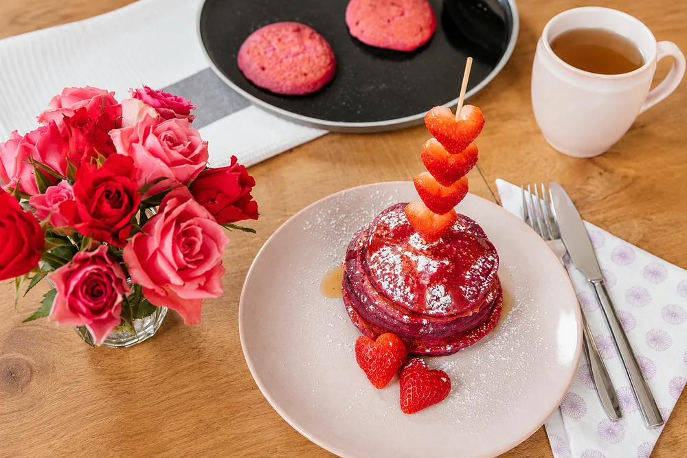 Rosa Pancakes mit Erdbeersauce, herzförmig geschnittenen Erdbeeren, Rosen und Tee auf einem Holztisch