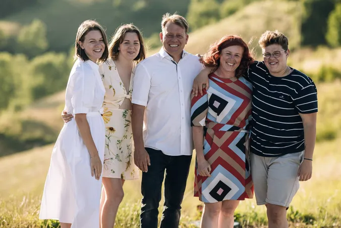 Eine Gruppe von fünf lächelnden Menschen, die eng beieinander auf einem sonnigen Feld stehen, im Hintergrund ist Grün zu sehen.