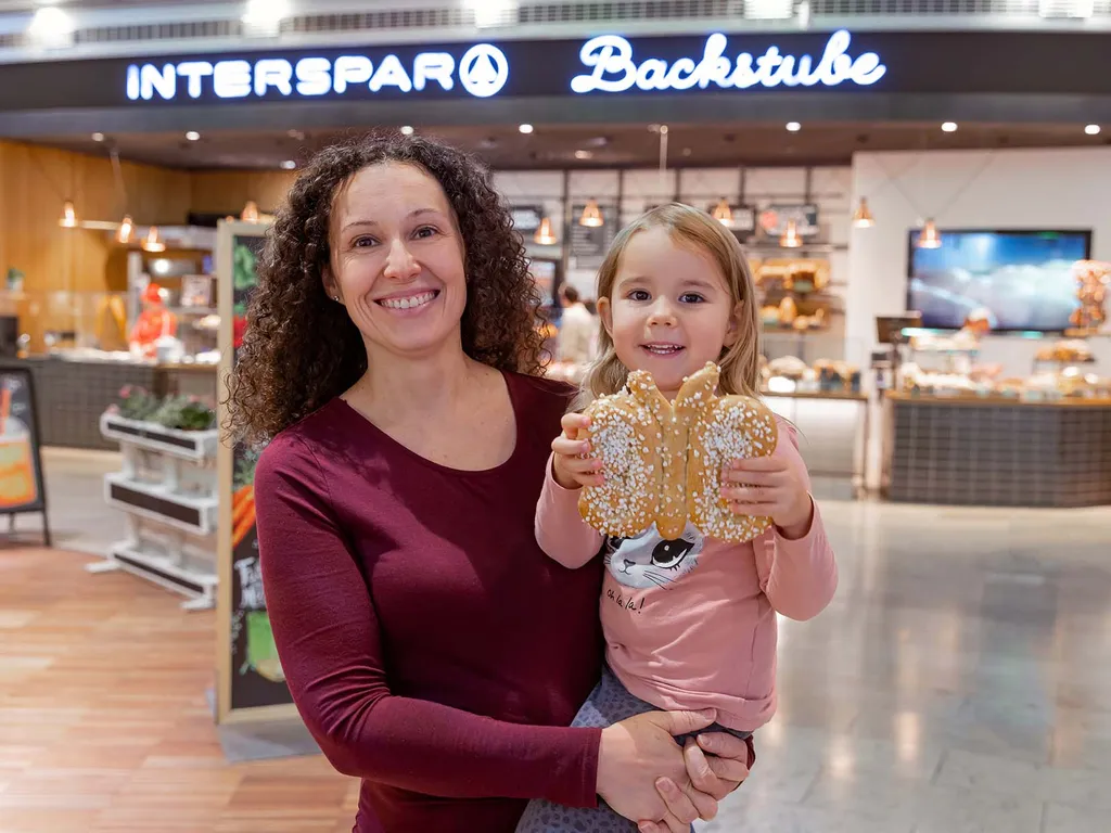 Eine lächelnde Frau mit einem kleinen Mädchen im Arm das grinsend einen Brioche-Schmetterling hält