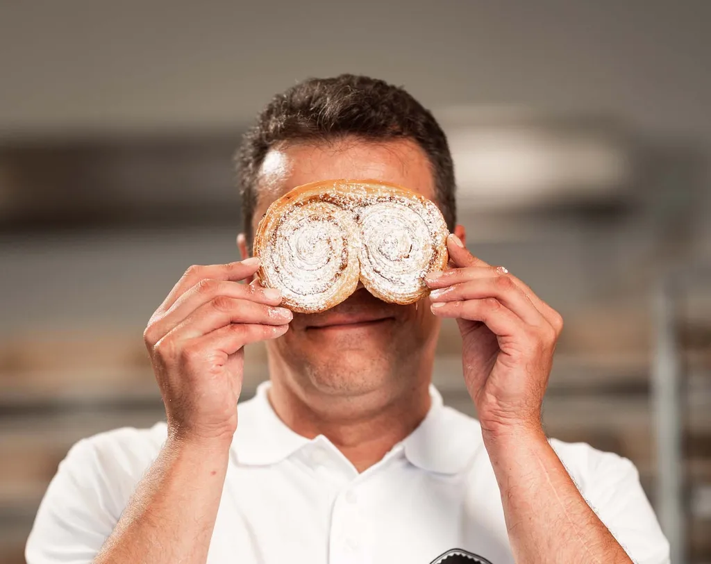 Ein INTERSPAR-Bäcker hält sich in einer Bäckerei spielerisch zwei mit Puderzucker bestäubte Gebäckstücke vor die Augen und lächelt.