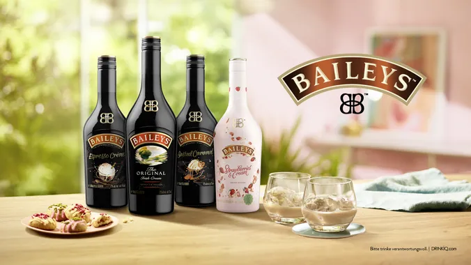 Vier Flaschen Baileys und zwei Gläser Sahnelikör auf einem Holztisch mit Gebäck und einem verschwommenen Garten im Hintergrund.