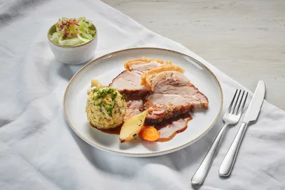 Ein Teller mit Schweinsbraten in Scheiben, Bratensoße, Knödel und Gemüse. Dazu gibt es Salat in einer Schüssel. Silberbesteck liegt neben dem Teller.
