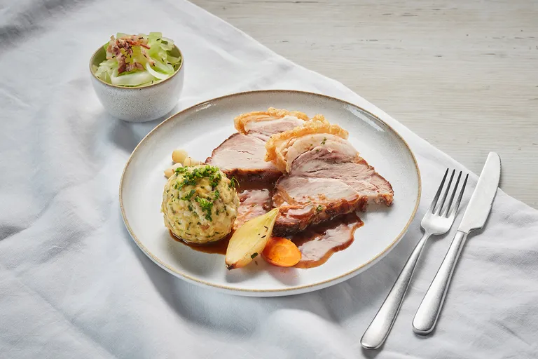 Ein Teller mit Schweinsbraten in Scheiben, Bratensoße, Knödel und Gemüse. Dazu gibt es Salat in einer Schüssel. Silberbesteck liegt neben dem Teller.