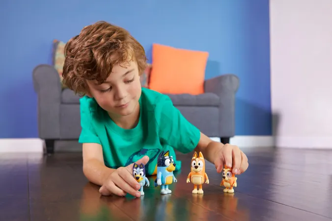 Kind liegt am Boden im Wohnzimmer, 2 Spielfiguren in der Hand, 2 am Boden, lächelnd