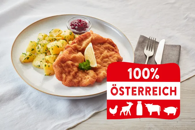Teller mit Schnitzel, Kartoffeln und Preiselbeeren. „100 % Österreich“-Etikett mit Symbolen. Serviette und Besteck neben dem Teller.