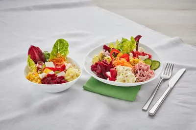 Zwei weiße Schüsseln mit buntem Salat, darunter Rote Bete, Kopfsalat, Mais und Schinken, auf einer weißen Tischdecke neben einer grünen Serviette und Besteck.