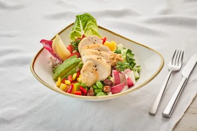 Eine Schüssel gemischter Salat mit Hähnchenstreifen, Avocado, Radieschen, Edamame und buntem Gemüse auf einer weißen Tischdecke, daneben das Besteck.