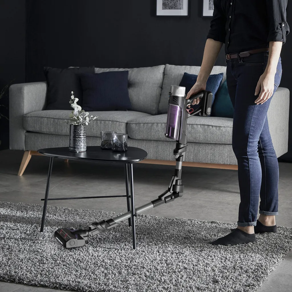Frau benutzt Akkustaubsauger von Rowenta mit flexiblem Saugrohr, saugt unter  schwarzem Couchtisch,  darunter ein grauer Teppich und im Hintergrund eine graue Couch mit Holzbeinen