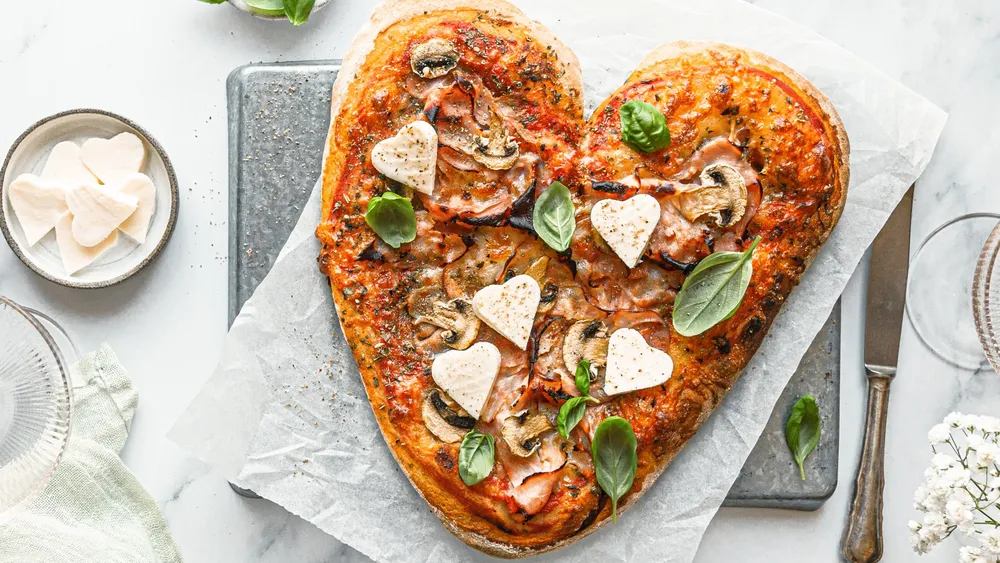 herzförmige Pizza auf einem hellen Tisch mit Backpapier und Steinbrett