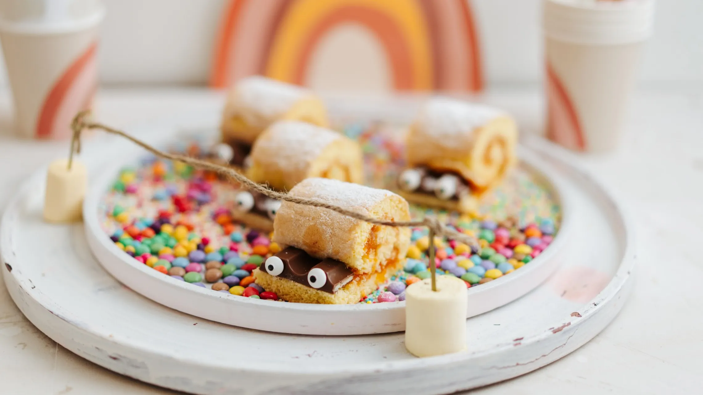 Bunte Biskuitrollen mit Zuckeraugen auf einem Tablett mit Streuseln, verziert mit Marshmallow-Schnüren, Tassen und einem Regenbogenhintergrund.