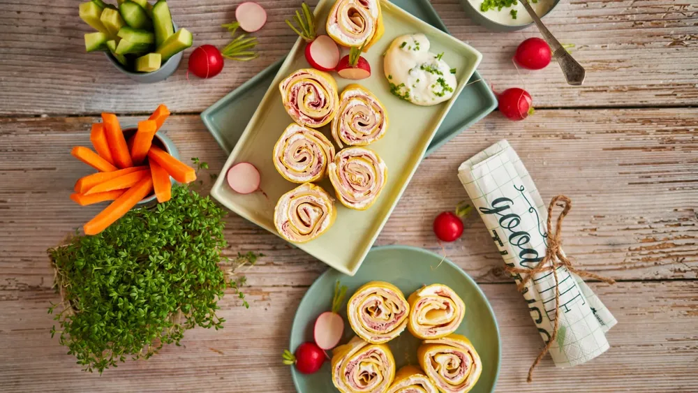 Schinken-Käse-Röllchen auf Tellern, garniert mit Radieschen und Dip, umgeben von Karotten- und Gurkenstangen.
